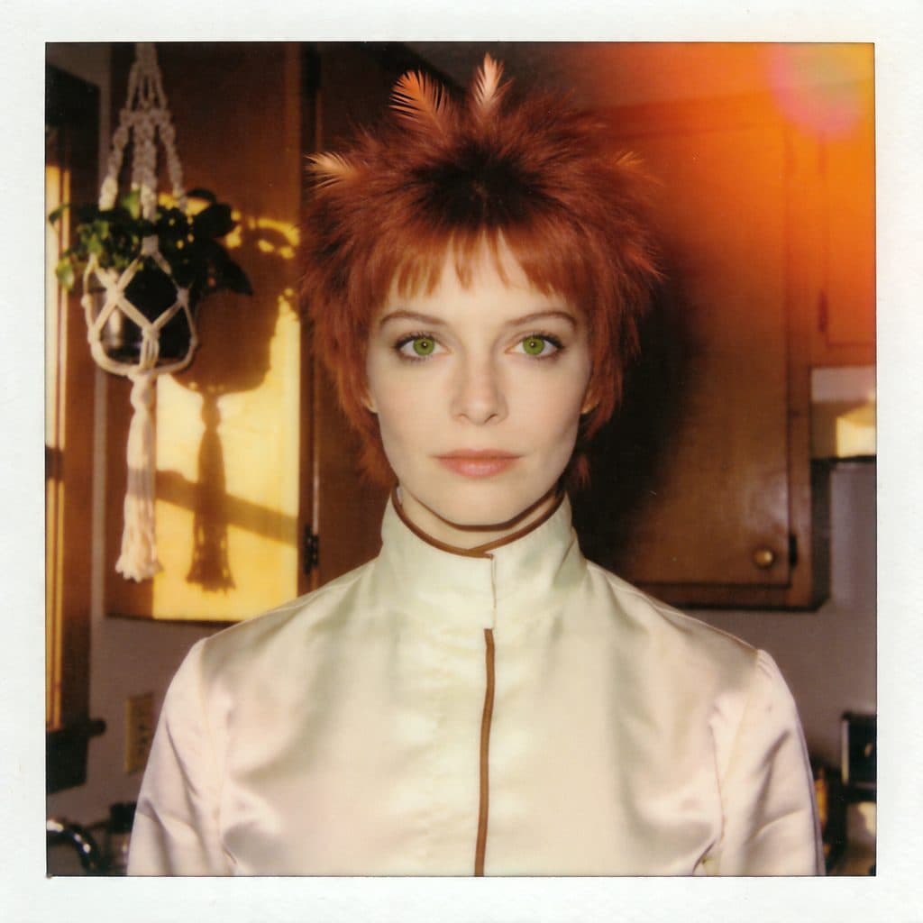 A faded 1974 Polaroid SX-70 photo of copper-haired Mira in a cream blouse, golden-hour kitchen setting.